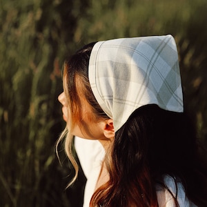 May include: A woman with long brown hair is wearing a white and green plaid bandana tied around her head. She is standing in a field of tall green grass.