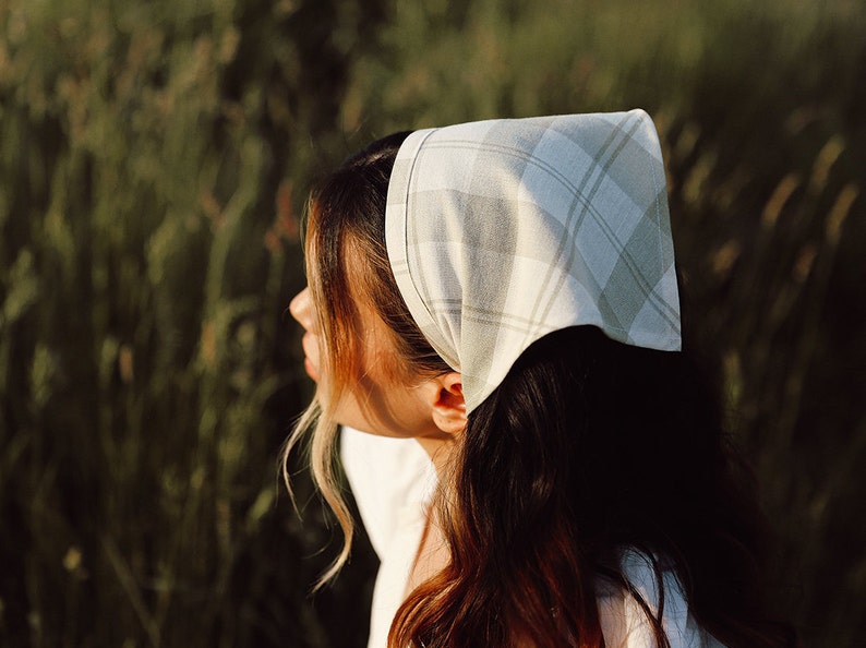 May include: A woman with long brown hair is wearing a white and green plaid bandana tied around her head. She is standing in a field of tall green grass.