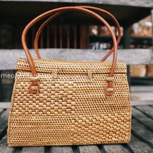 May include: A woven brown rattan handbag with leather handles and a flap closure. The bag is sitting on a wooden surface.