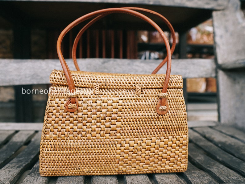 May include: A woven brown rattan handbag with leather handles and a flap closure. The bag is sitting on a wooden surface.