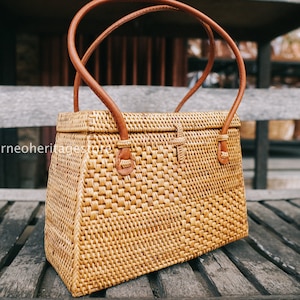 May include: A woven rattan handbag with brown leather handles and a rectangular shape. The bag is sitting on a wooden surface.