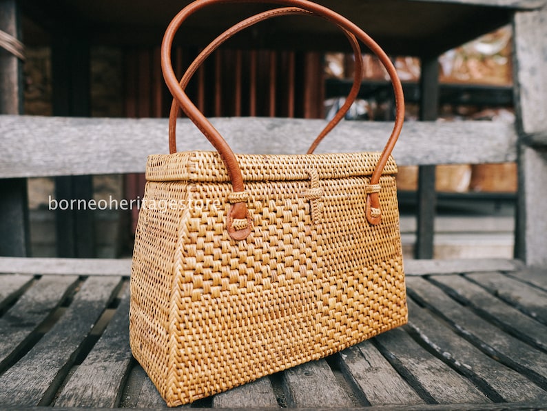 May include: A woven rattan handbag with brown leather handles and a rectangular shape. The bag is sitting on a wooden surface.