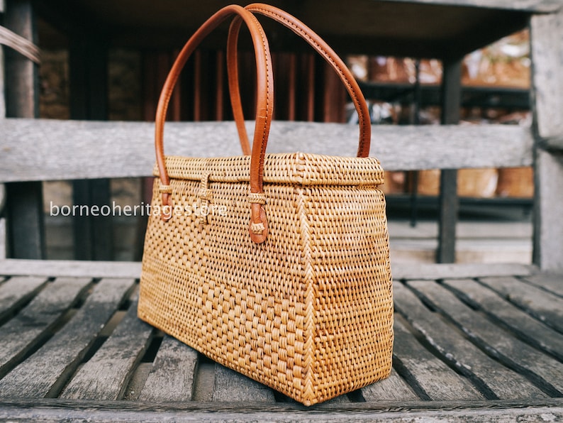 May include: A woven rattan handbag with brown leather handles. The bag has a rectangular shape and a woven pattern.