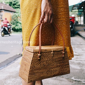 May include: A brown woven handbag with a rectangular shape and a brown leather handle. The bag is made of natural materials and has a woven texture.