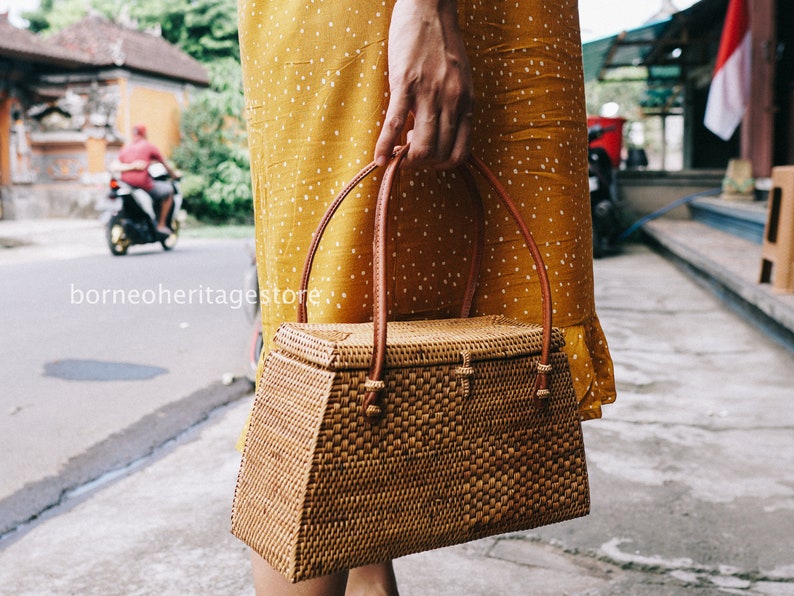 May include: A brown woven handbag with a rectangular shape and a brown leather handle. The bag is made of natural materials and has a woven texture.