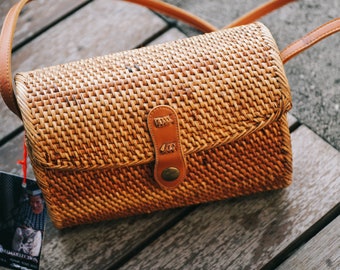 May include: A brown woven crossbody bag with a leather strap and a brown button closure. The bag is made of natural materials and has a rustic look.