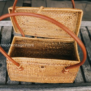 May include: A woven rattan box with a brown leather handle. The box is open and the inside is visible. The box is sitting on a wooden surface.