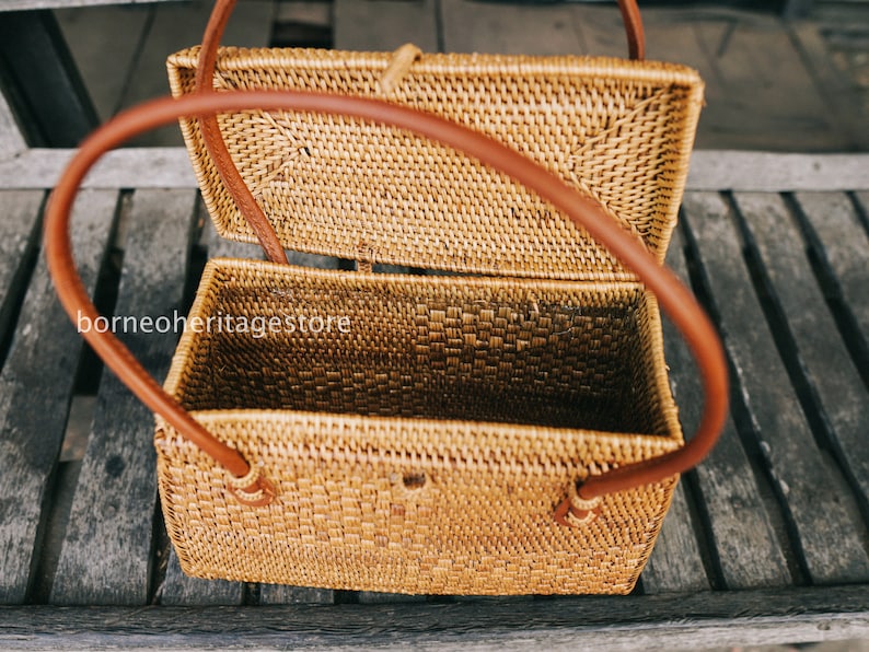 May include: A woven rattan box with a brown leather handle. The box is open and the inside is visible. The box is sitting on a wooden surface.