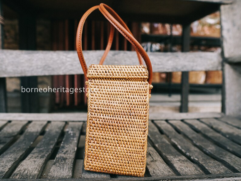 May include: A brown woven rattan handbag with a brown leather strap. The bag is sitting on a wooden surface.