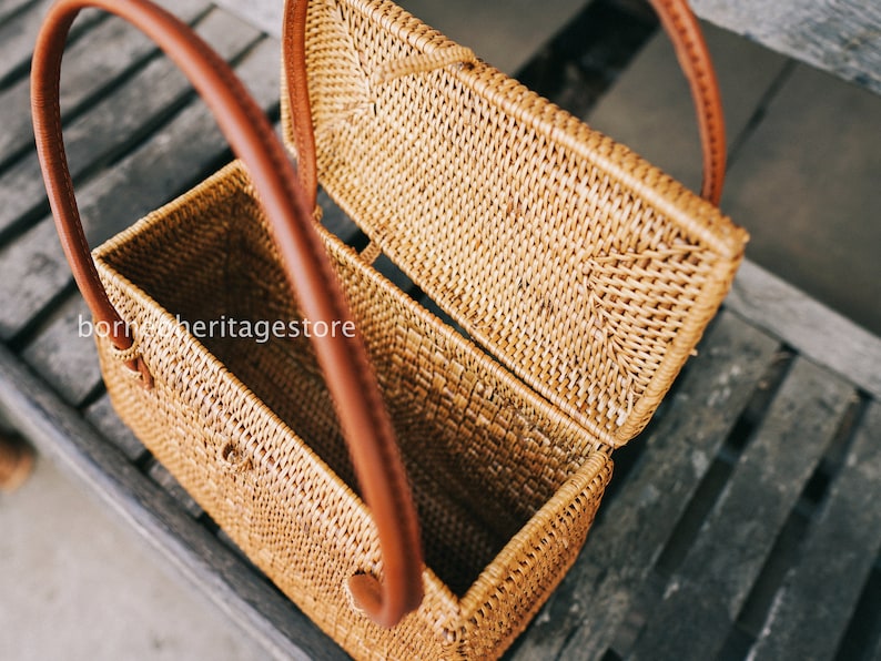 May include: A woven rattan box with a hinged lid and two brown leather handles. The box is open and the inside is visible.