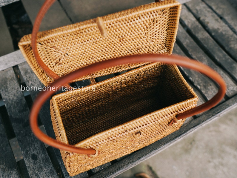 May include: A woven rattan handbag with a brown leather handle. The bag is open and the inside is visible. The bag is sitting on a wooden surface.