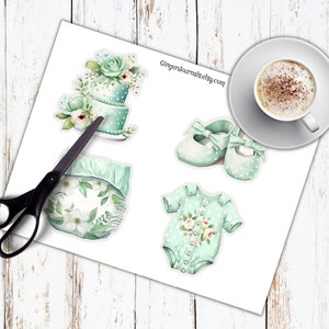 May include: A sheet of stickers featuring baby-themed illustrations. Includes a two-tiered cake, a diaper, baby shoes with bows, and a onesie, all in shades of mint green and white. A pair of scissors is next to the stickers. A cup of coffee sits on a saucer.