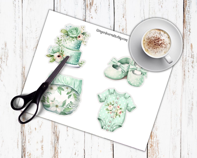 May include: A sheet of stickers featuring baby-themed illustrations. Includes a two-tiered cake, a diaper, baby shoes with bows, and a onesie, all in shades of mint green and white. A pair of scissors is next to the stickers. A cup of coffee sits on a saucer.