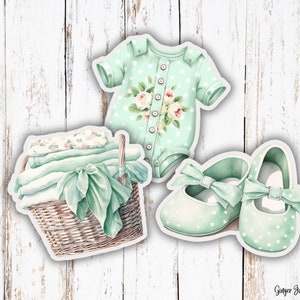 May include: Watercolor illustration of baby items: a mint green onesie with floral accents, a pair of polka-dot shoes with bows, and a wicker basket with folded blankets and a ribbon. The items are on a white wooden background.