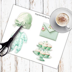 May include: A sheet of paper with watercolor illustrations of baby items, including a green hat with a bow, a onesie, an envelope with roses, and cupcakes on a stand. A cup of coffee sits on a saucer. Scissors are also present.