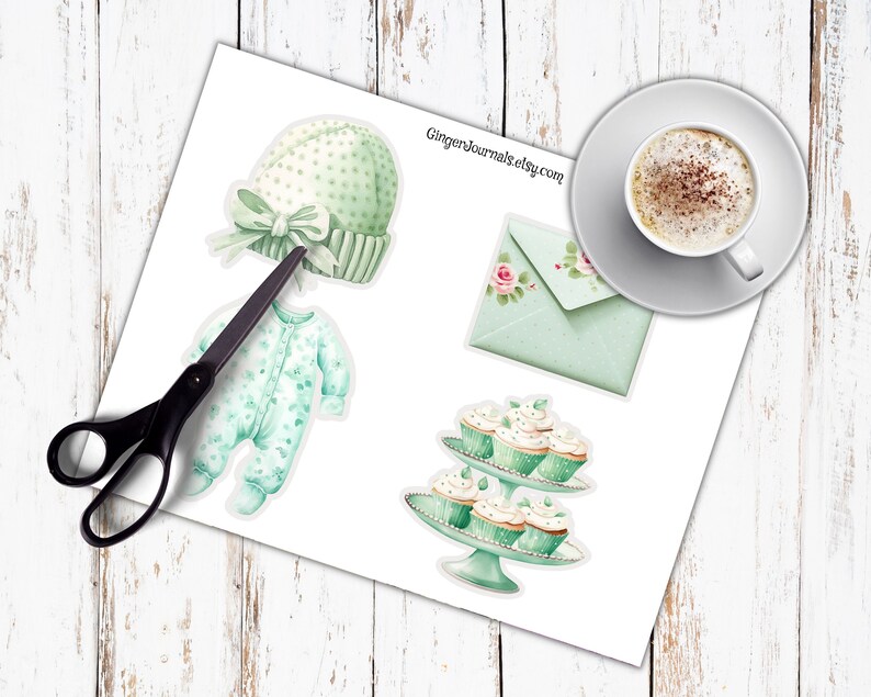 May include: A sheet of paper with watercolor illustrations of baby items, including a green hat with a bow, a onesie, an envelope with roses, and cupcakes on a stand. A cup of coffee sits on a saucer. Scissors are also present.