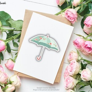 May include: A sticker of a teal umbrella with a floral pattern and polka dots. The umbrella has a red handle and is set on a white card with a brown envelope. Pink roses surround the card. The text "Ginger Journals" is in the top left corner.