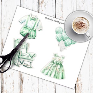 May include: Watercolor stickers of baby-themed items, including a green romper with suspenders, a rocking chair, a dress, and balloons. A cup of coffee sits on a saucer in the upper right corner. The text "GingerJournals.etsy.com" is visible.