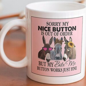May include: White ceramic mug with a pink background and a humorous saying: "Sorry my nice button is out of order but my bite me button works just fine". Three cartoon donkeys with pink sunglasses, a black and white bandana, and a sunflower are featured on the mug.