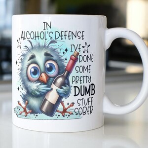 May include: White ceramic mug with a blue bird illustration holding a bottle of wine. The mug has the text "In alcohol's defense I've done some pretty dumb stuff sober."