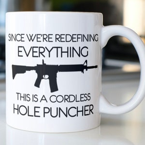 May include: White ceramic mug with a black silhouette of a rifle and the text "Since we're redefining everything, this is a cordless hole puncher."