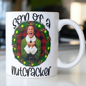 May include: White ceramic mug with a green, red, and black Christmas wreath design featuring Buddy the Elf from the movie Elf. The text "son of a nutcracker" is printed on the mug.