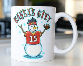 May include: White ceramic mug with a snowman wearing a red jersey with the number 15 and a football in its hand. The mug has the text "Kansas City" with stars around it.