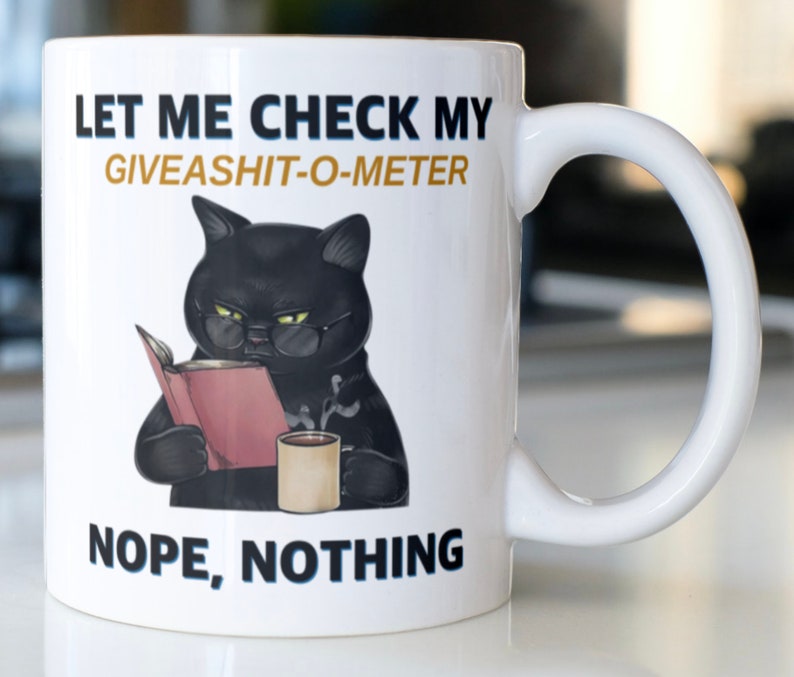 May include: White ceramic mug with a black cat wearing glasses and reading a book. The mug has the text "Let me check my giveashit-o-meter. Nope, nothing."