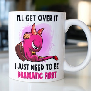 May include: White ceramic mug with a pink dragon illustration and the text "I'll get over it I just need to be dramatic first".