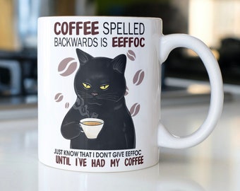 May include: White ceramic coffee mug with a black cat illustration holding a cup of coffee. The mug has the text "COFFEE SPELLED BACKWARDS IS EEFFOC JUST KNOW THAT I DON'T GIVE EEFFOC UNTIL I'VE HAD MY COFFEE".