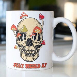 May include: White ceramic mug with a skull design and mushrooms growing out of the eye sockets. The text "STAY WEIRD AF" is printed in red, yellow, and green letters on the bottom of the mug.