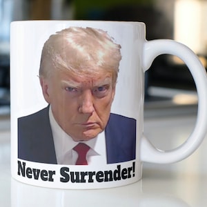 May include: White ceramic coffee mug with a color image of a man with blonde hair and a serious expression. The text "Never Surrender!" is printed in black at the bottom of the mug.