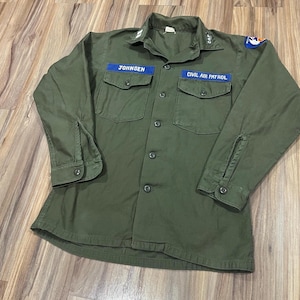Op de afbeelding: Olijfgroen Civil Air Patrol uniformshirt met knoopsluiting, twee borstzakken en lange mouwen. Voorzien van een blauw naamlabel met "JOHNSEN" en een "CIVIL AIR PATROL" patch.