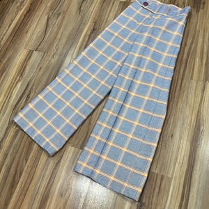 May include: Light blue plaid pants with a high waist and wide legs. The plaid pattern features yellow and red lines over a light blue background. The pants have a button closure at the waist.