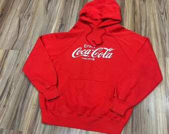 May include: A red hooded sweatshirt with a front pocket and white "Coca-Cola" logo. The sweatshirt has a drawstring hood and long sleeves. The text "Enjoy" is above the Coca-Cola logo.