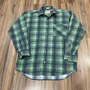 May include: Green and navy plaid button-down shirt with a chest pocket. The long-sleeved shirt has a collared neckline and buttoned cuffs. The shirt is made of a soft, textured fabric and is laid flat on a wooden surface.