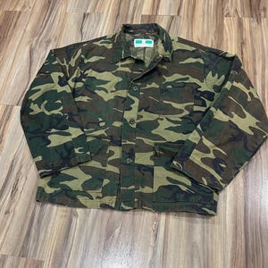 May include: A long-sleeve button-up jacket featuring a camouflage pattern in shades of dark green, brown, and black. The jacket includes a pointed collar, a single chest pocket on the left side, two lower front pockets, and a white interior label.