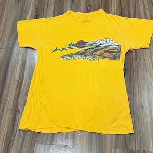 Peut inclure: T-shirt jaune moutarde à col montant. Le devant présente un graphisme de coucher de soleil, de montagnes et d'oiseaux, avec le texte "PACIFIC UNIVERSITY" en bleu.