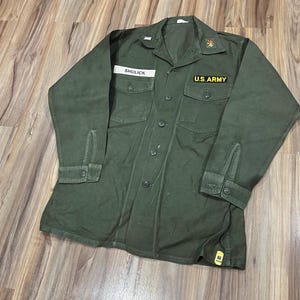 Può includere: Una camicia militare verde oliva con il nome "SHULICK" su un'etichetta sopra la tasca sinistra. La tasca destra ha una toppa "U.S. ARMY". La camicia ha una chiusura con bottoni, due tasche sul petto e maniche lunghe.