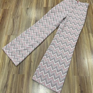 May include: A pair of patterned pants with a white background and a repeating zigzag design in gray and red. The pants are laid out on a wooden surface, showcasing the full length and pattern.