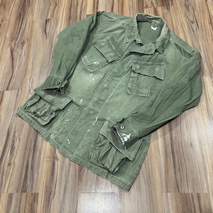 Può includere: Giacca verde oliva in stile militare con più tasche e chiusure con bottoni. La giacca ha maniche lunghe e un colletto. Schizzi di vernice bianca sono visibili sul davanti e sulle maniche. La giacca è appoggiata su una superficie di legno.