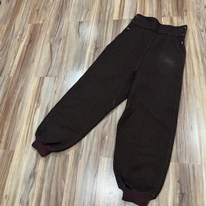 May include: Dark brown high-waisted pants featuring a wide waistband with two buttons on each side, a center seam down the front of each leg, and ribbed knit cuffs at the ankles. The pants have a structured appearance with a solid dark brown color.