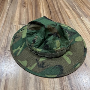 May include: A camouflage boonie hat featuring a wide brim, a rounded crown, and two black ventilation eyelets on each side. The hat is constructed from a durable fabric with a green, brown, and black woodland pattern and includes a fabric band.