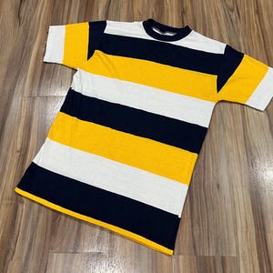 Może przedstawiać: T-shirt z krótkim rękawem i okrągłym dekoltem, ozdobiony poziomymi pasami w kolorach granatowym, jaskrawożółtym i białym. Odzież posiada jednolity granatowy kołnierzyk oraz proste wykończenie dołu z konsekwentnym wzorem w paski na całej powierzchni.