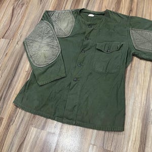 Könnte beinhalten: Ein olivgrünes Hemd im Militärstil mit rundem Kragen und Knopfleiste. Das Hemd hat zwei Brusttaschen und verstärkte Aufnäher an Schultern und Ellbogen. Die Aufnäher sind in einem helleren Grünton gehalten und haben ein Stichdesign.