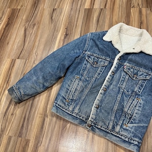 May include: A blue denim jacket with a sherpa lining. The jacket has a button closure, two front pockets, and a single pocket on the left chest.