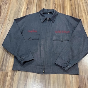 May include: A charcoal gray work jacket featuring a full-length front zipper, a pointed collar, and long sleeves with buttoned cuffs. The chest displays two patch pockets with buttoned flaps, with the word Captain and the text Hotel Plaza embroidered in red.
