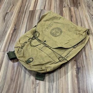 May include: A tan canvas backpack featuring a large flap closure with a drawstring cord. The flap displays the text "PAINE" and a circular Boy Scouts of America emblem. The bag includes dark green webbing straps, metal D-rings, and metal grommets.