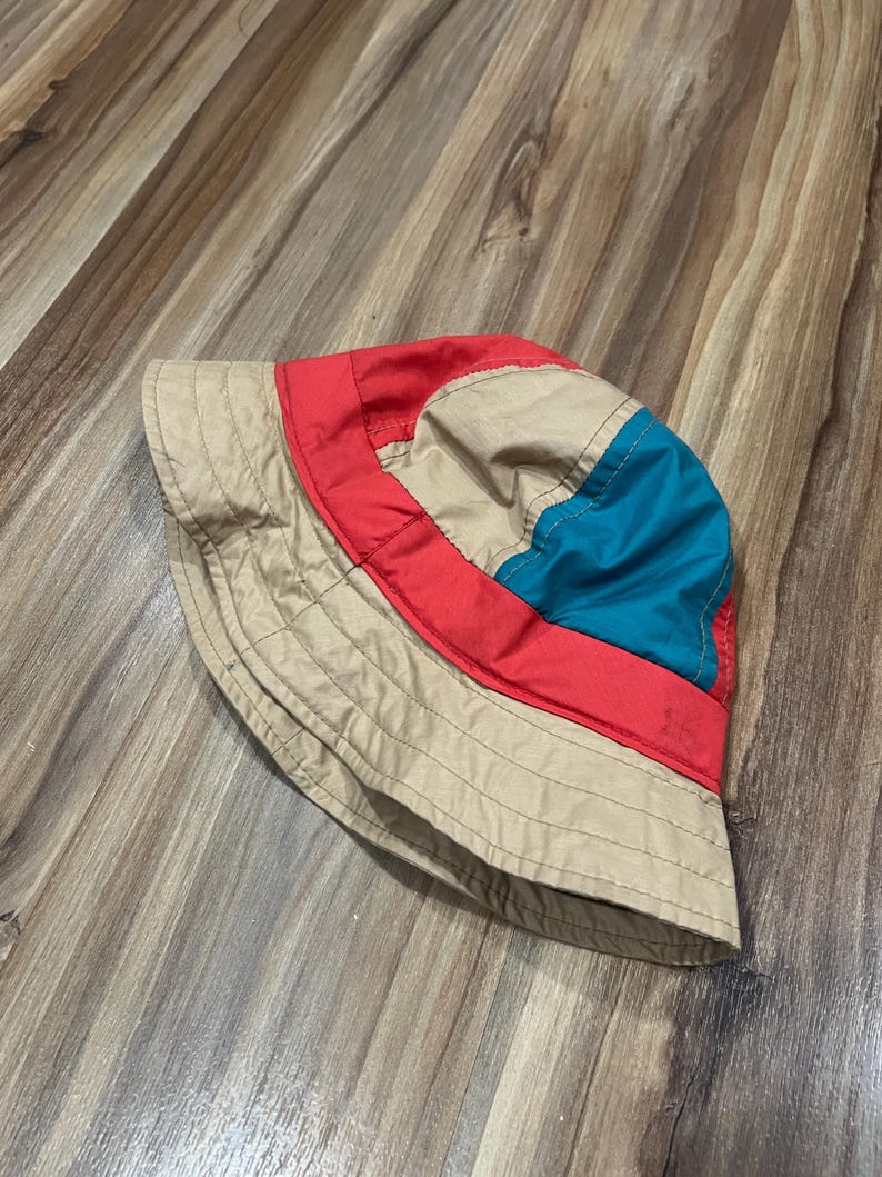 May include: A bucket hat featuring a color-blocked design with a wide beige brim and crown panels. The hat incorporates a vibrant red band encircling the midsection and a distinct teal blue panel on the upper side, all constructed with visible stitching.
