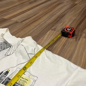 May include: A white t-shirt with a graphic print of medical equipment and text. A yellow measuring tape extends across the shirt, showing measurements in inches. The tape measure is black and red.